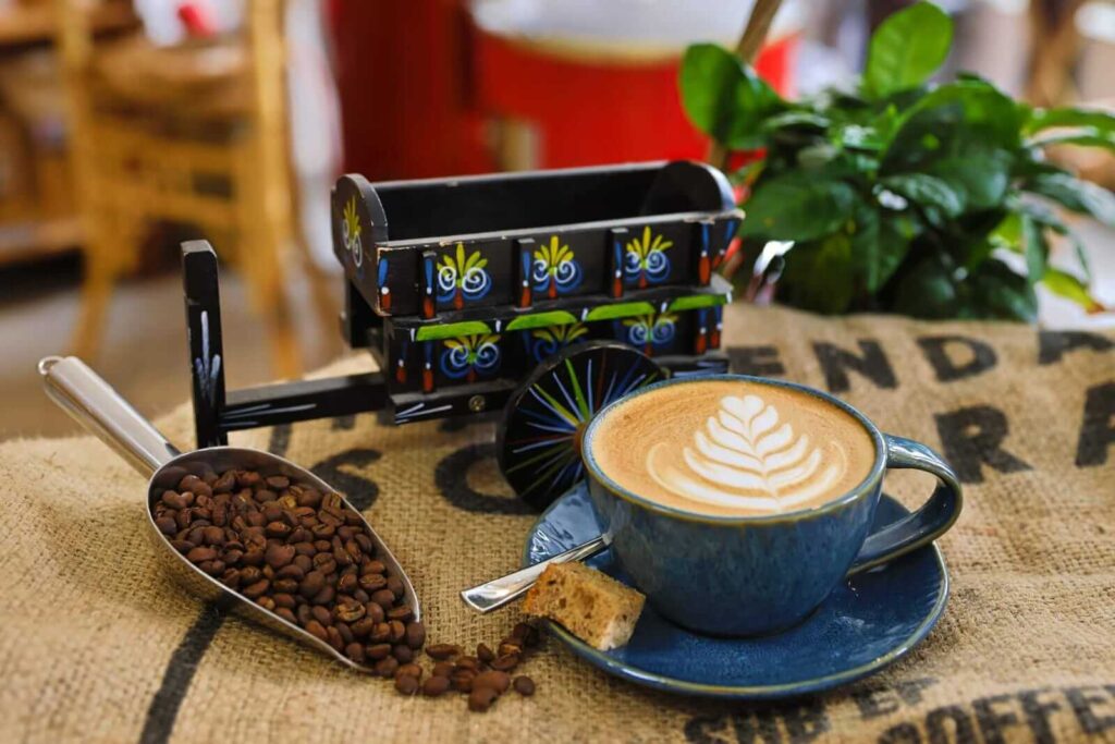 Frisch gebrühter Cappuccino mit Latte Art, daneben Kaffeebohnen und eine bunte Mini-Kaffeekarre.